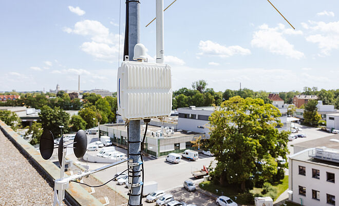 Auf einem Dach ist ein technisches Mess- und Kommunikationsgerät an einem Mast befestigt. Das weiße Gehäuse enthält Antennen und Kabel, darunter ist ein Windmessgerät mit zwei Schalen und einem Windrichtungsanzeiger montiert. Im Hintergrund sind Gebäude, Bäume und parkende Fahrzeuge zu sehen, bei klarem Himmel mit vereinzelten Wolken. Verlinkung zur IoT Seite.