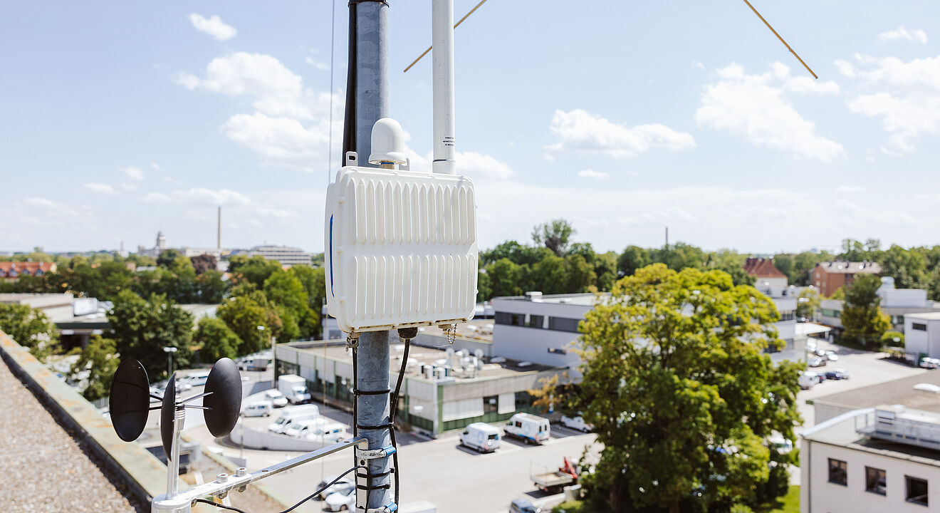 Auf einem Dach ist ein technisches Mess- und Kommunikationsgerät an einem Mast befestigt. Das weiße Gehäuse enthält Antennen und Kabel, darunter ist ein Windmessgerät mit zwei Schalen und einem Windrichtungsanzeiger montiert. Im Hintergrund sind Gebäude, Bäume und parkende Fahrzeuge zu sehen, bei klarem Himmel mit vereinzelten Wolken. Verlinkung zur IoT Seite.