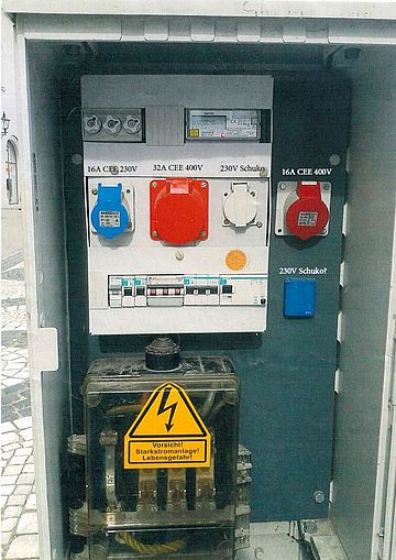 Geöffneter mobiler Stromverteiler im Außenbereich mit integriertem Stromzähler, Sicherungen und mehreren Industrie-Steckdosen. Sichtbar sind eine blaue 16-A-CEE-Steckdose (230 V), eine rote 32-A-CEE-Steckdose (400 V), eine weiße Schuko-Steckdose (230 V) sowie eine weitere rote 16-A-CEE-Steckdose (400 V). Im unteren Bereich befindet sich ein transparenter Anschlusskasten mit deutlich sichtbarem gelbem Warnschild „Vorsicht! Starkstromanlage! Lebensgefahr!“. Die Anlage dient der sicheren temporären Stromversorgung bei Veranstaltungen, Baustellen oder Infrastrukturmaßnahmen.