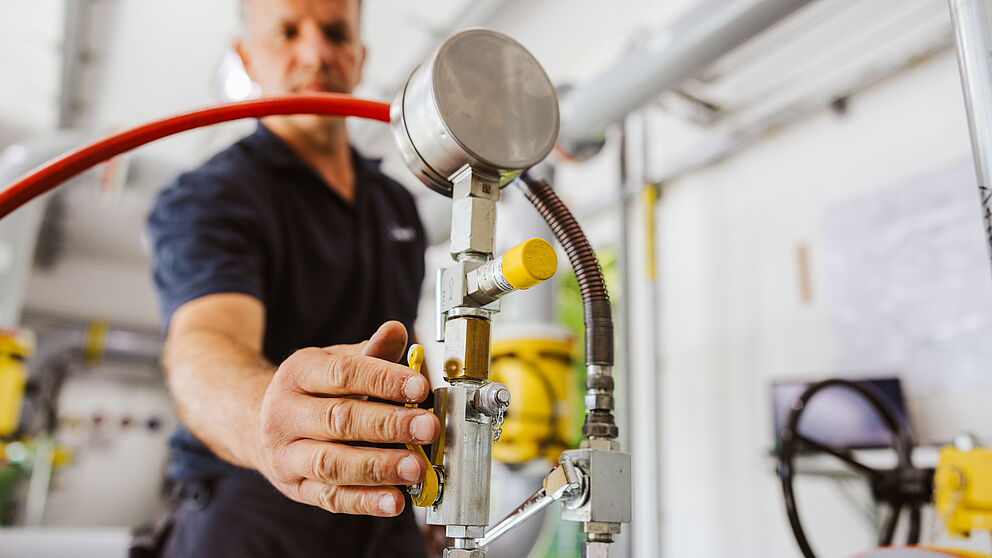 Nahaufnahme einer Hand, die ein Ventil an einer Rohrleitungsanlage bedient. Sichtbar sind ein Manometer, metallische Leitungen, gelbe Armaturen und ein rotes Schlauchsystem. Im Hintergrund befinden sich weitere Rohre und technische Geräte in einem hellen Raum. Verlinkung zur Gasanalyse Seite.