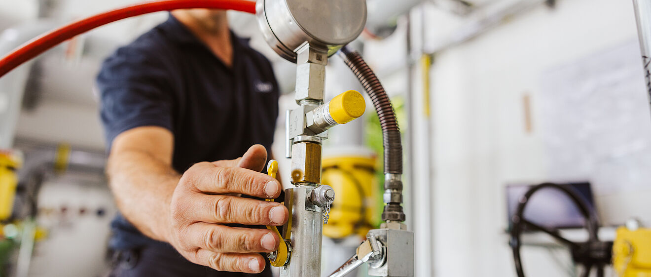 Nahaufnahme einer Hand, die ein Ventil an einer Rohrleitungsanlage bedient. Sichtbar sind ein Manometer, metallische Leitungen, gelbe Armaturen und ein rotes Schlauchsystem. Im Hintergrund befinden sich weitere Rohre und technische Geräte in einem hellen Raum. Verlinkung zur Gasanalyse Seite.