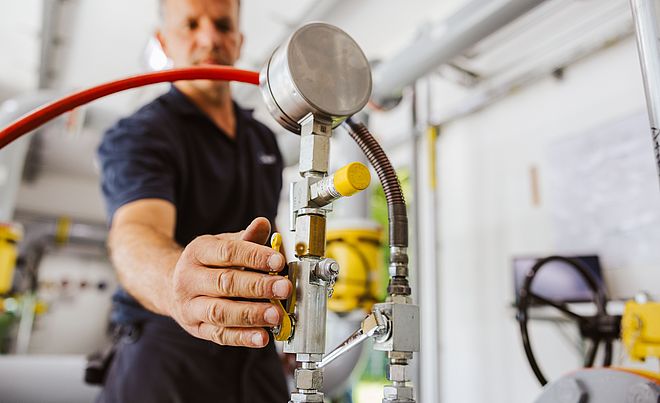 Nahaufnahme einer Hand, die ein Ventil an einer Rohrleitungsanlage bedient. Sichtbar sind ein Manometer, metallische Leitungen, gelbe Armaturen und ein rotes Schlauchsystem. Im Hintergrund befinden sich weitere Rohre und technische Geräte in einem hellen Raum. Verlinkung zur Gasanalyse Seite.