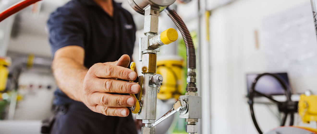 Nahaufnahme einer Hand, die ein Ventil an einer Rohrleitungsanlage bedient. Sichtbar sind ein Manometer, metallische Leitungen, gelbe Armaturen und ein rotes Schlauchsystem. Im Hintergrund befinden sich weitere Rohre und technische Geräte in einem hellen Raum. Verlinkung zur Gasanalyse Seite.