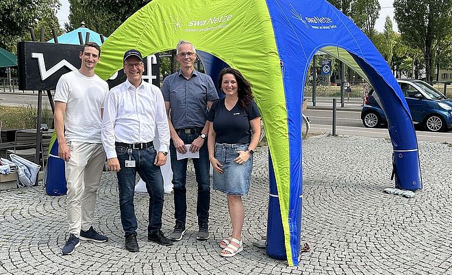 Vier Personen stehen unter einem grün-blauen Informationszelt der swa Netze auf einem gepflasterten Platz im öffentlichen Raum. Auf dem Zelt ist der Schriftzug „swa Netze – Immer an deiner Seite“ sichtbar. Die Gruppe präsentiert sich vor einem Infostand im Rahmen einer Veranstaltung zur Bürger- und Kundenkommunikation. Im Hintergrund sind Straßenraum, Bäume, Fahrzeuge und weitere Veranstaltungselemente zu sehen. Die Aufnahme vermittelt die Präsenz und Ansprechbarkeit von swa Netze vor Ort.