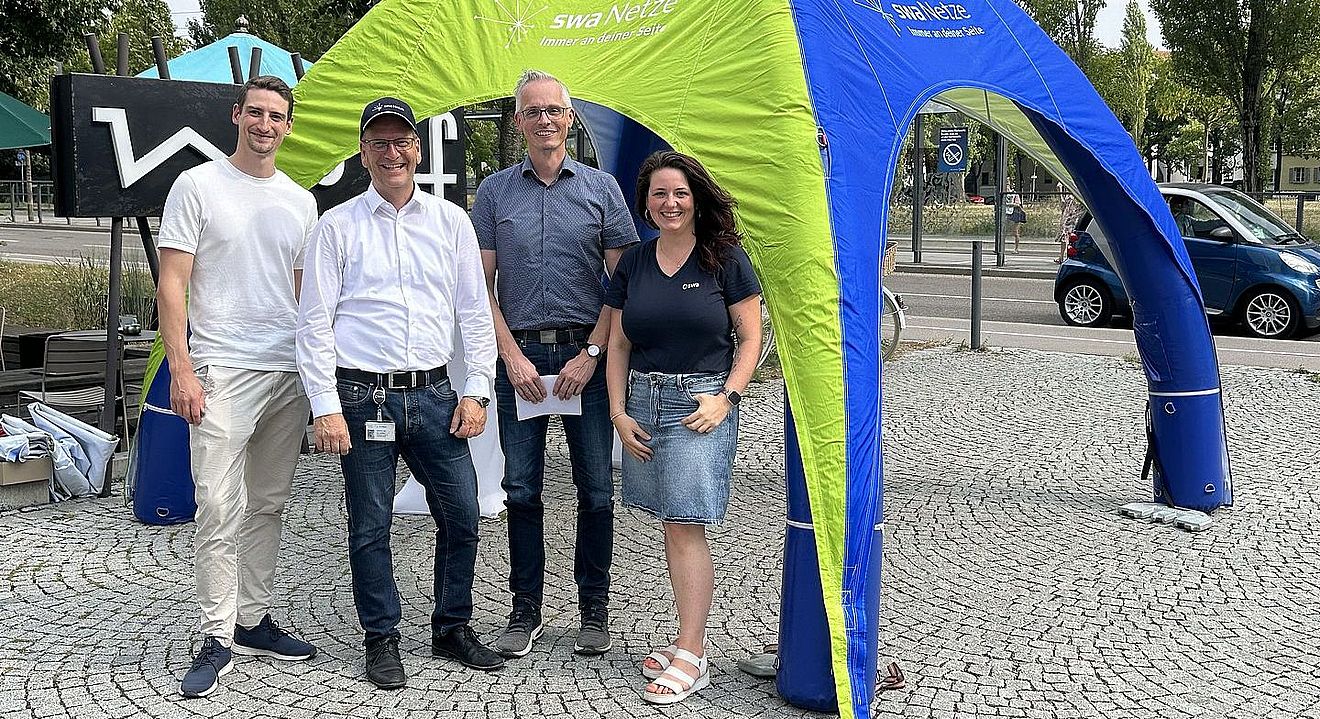 Vier Personen stehen unter einem grün-blauen Informationszelt der swa Netze auf einem gepflasterten Platz im öffentlichen Raum. Auf dem Zelt ist der Schriftzug „swa Netze – Immer an deiner Seite“ sichtbar. Die Gruppe präsentiert sich vor einem Infostand im Rahmen einer Veranstaltung zur Bürger- und Kundenkommunikation. Im Hintergrund sind Straßenraum, Bäume, Fahrzeuge und weitere Veranstaltungselemente zu sehen. Die Aufnahme vermittelt die Präsenz und Ansprechbarkeit von swa Netze vor Ort.