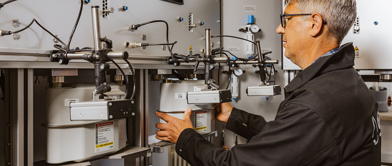 Eine Person arbeitet an einer Prüfanlage für Gaszähler. Mehrere Gaszähler sind in einer Reihe montiert und über Leitungen sowie Messgeräte miteinander verbunden. Die Anlage besteht aus grauen Metallplatten mit Anzeigen und Anschlüssen. Die Person hält einen Gaszähler, der in die Vorrichtung eingesetzt wird. Verlinkung zur Messstellenbetrieb Gas Seite.