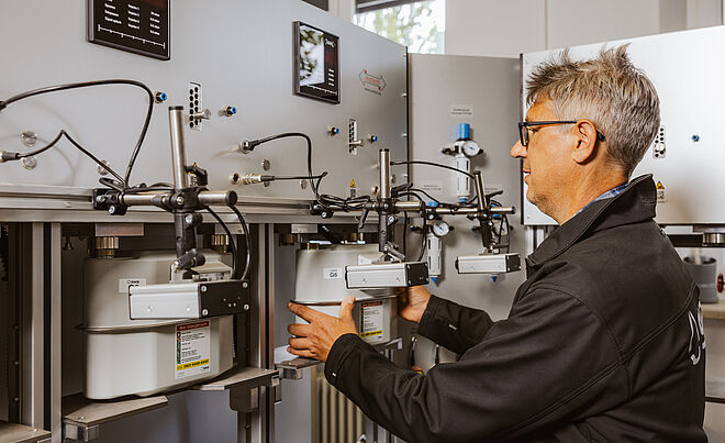 Eine Person arbeitet an einer Prüfanlage für Gaszähler. Mehrere Gaszähler sind in einer Reihe montiert und über Leitungen sowie Messgeräte miteinander verbunden. Die Anlage besteht aus grauen Metallplatten mit Anzeigen und Anschlüssen. Die Person hält einen Gaszähler, der in die Vorrichtung eingesetzt wird. Verlinkung zur Messstellenbetrieb Gas Seite.