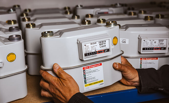 Mehrere Gaszähler sind in einem Regal aufgereiht. Im Vordergrund hält eine Person einen einzelnen Gaszähler mit beiden Händen. Der Zähler ist grau, mit einem Display und einem Aufkleber, der Hinweise für den Notfall bei Gasgeruch enthält sowie die Telefonnummer des Störungsdienstes. Auf den Geräten sind Typenbezeichnungen wie ‚BK-G4M‘ und ‚G6‘ sowie das Logo der swa Netze sichtbar. Verlinkung zur Zählerwechsel Seite.