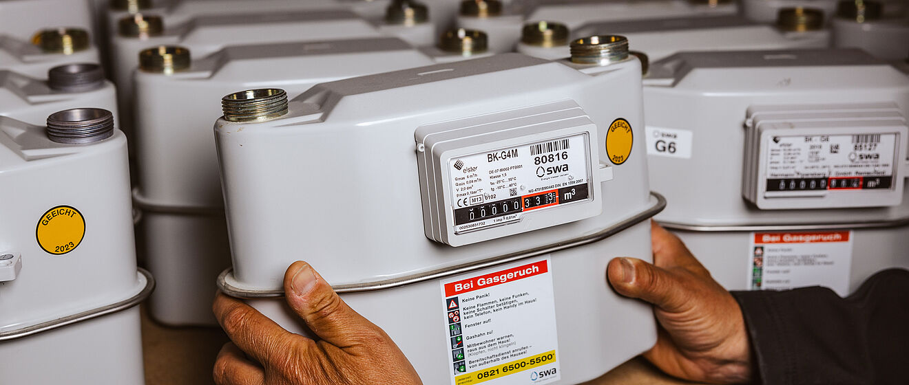 Mehrere Gaszähler sind in einem Regal aufgereiht. Im Vordergrund hält eine Person einen einzelnen Gaszähler mit beiden Händen. Der Zähler ist grau, mit einem Display und einem Aufkleber, der Hinweise für den Notfall bei Gasgeruch enthält sowie die Telefonnummer des Störungsdienstes. Auf den Geräten sind Typenbezeichnungen wie ‚BK-G4M‘ und ‚G6‘ sowie das Logo der swa Netze sichtbar. Verlinkung zur Zählerwechsel Seite.