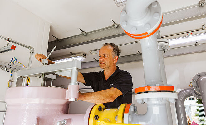 Technikraum mit Rohrleitungsanlage: Eine Person arbeitet an einem großen, hellrosa Druckbehälter, der mit grauen und gelben Rohrleitungen verbunden ist. Die Rohre sind mit roten Markierungen versehen, und im Hintergrund sind Kabel, Schaltkästen und Beleuchtung sichtbar. Verlinkung zur Einspeisung Gas Seite.