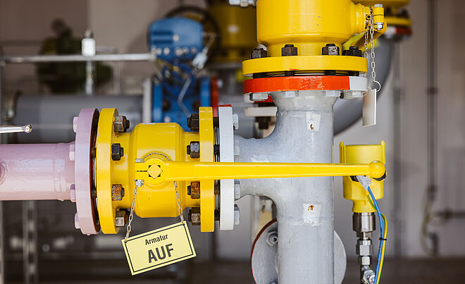 Detailaufnahme einer Rohrleitungsanlage mit gelber Armatur und Hebel, der auf ‚AUF‘ steht. Die Rohre sind grau und rosa, verbunden durch Flansche mit roten Markierungen. Im Hintergrund sind weitere technische Komponenten und Leitungen sichtbar.