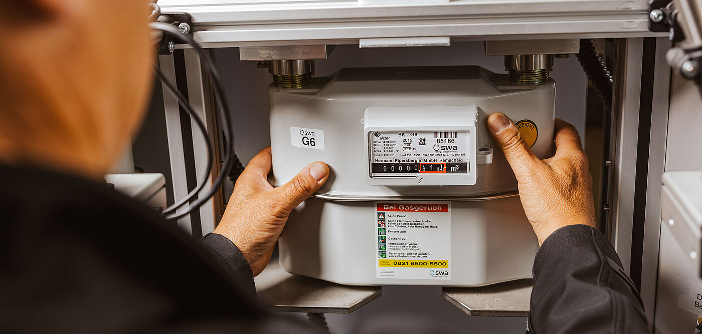 Eine Person setzt einen Gaszähler in eine Prüfvorrichtung ein. Der Gaszähler ist vom Typ G6 und zeigt ein Display mit Zählerstand sowie technischen Angaben. Auf dem Gerät befindet sich ein Aufkleber mit Warnhinweisen und Prüfinformationen. Die Prüfanlage besteht aus einer Metallkonstruktion mit Halterungen und einem QR-Code oben.