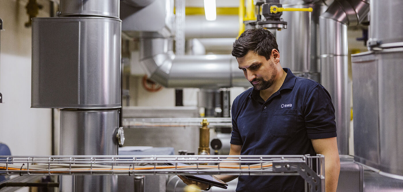 Technikraum mit großen silbernen Rohrleitungen, gelben Gasleitungen und Armaturen. Eine Person in dunkler Arbeitskleidung steht vor der Anlage und hält ein Tablet, offenbar zur Kontrolle oder Wartung der Installation. Im Hintergrund sind weitere Rohre und eine helle Deckenbeleuchtung sichtbar.