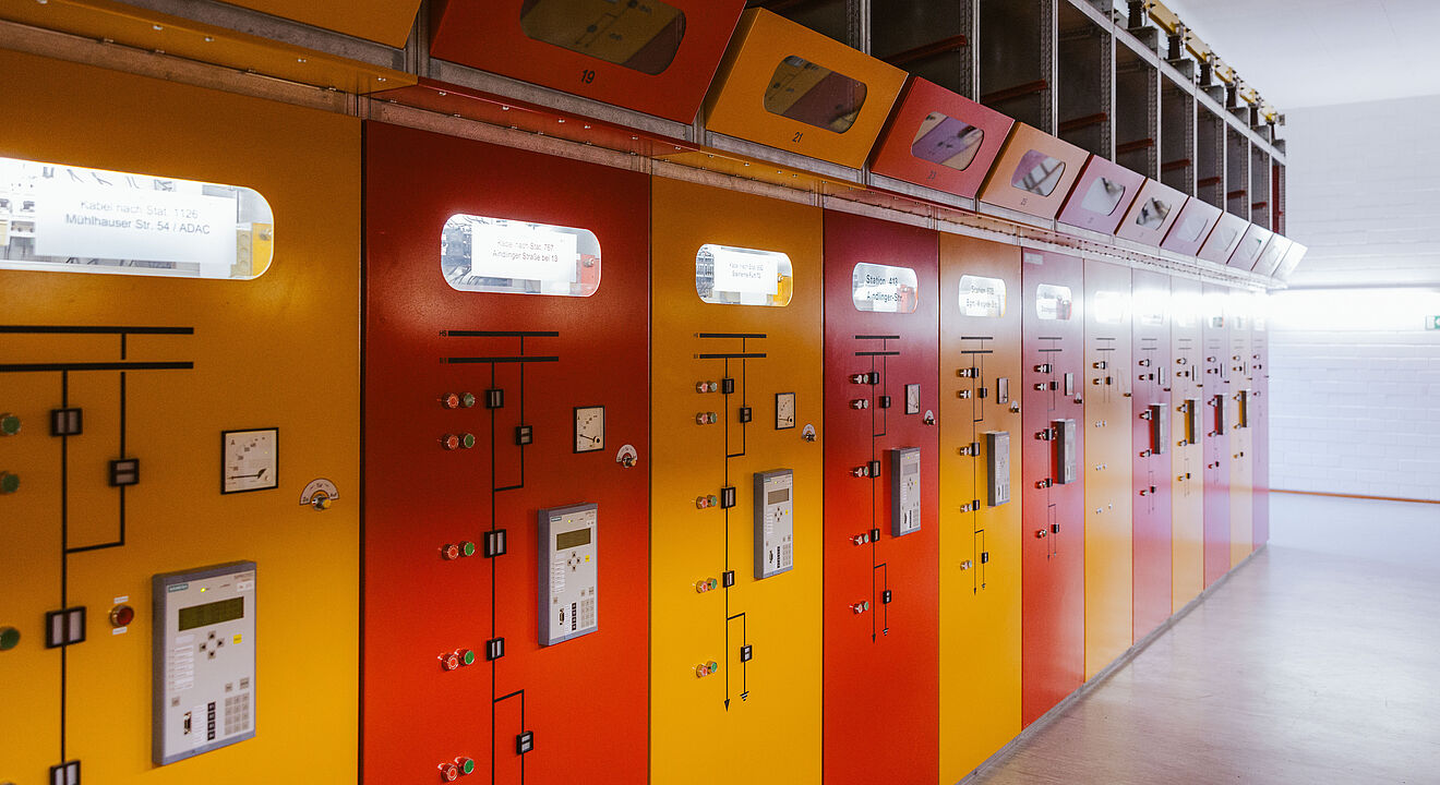 Reihe von farbigen Schaltanlagen in einem Technikraum: Die Fronten der Schaltschränke sind in Rot und Gelb gehalten und mit Anzeigen, Schaltern und Bedienelementen ausgestattet. Oberhalb der Schränke befinden sich Sichtfenster und Kabelkanäle. Die Szene zeigt eine moderne elektrische Schaltanlage für Energieverteilung und Netztechnik. Verlinkung zur Veröffentlichungen Strom Seite.