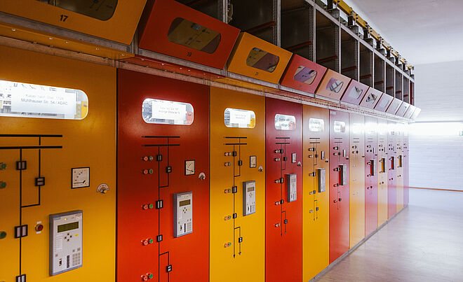 Reihe von farbigen Schaltanlagen in einem Technikraum: Die Fronten der Schaltschränke sind in Rot und Gelb gehalten und mit Anzeigen, Schaltern und Bedienelementen ausgestattet. Oberhalb der Schränke befinden sich Sichtfenster und Kabelkanäle. Die Szene zeigt eine moderne elektrische Schaltanlage für Energieverteilung und Netztechnik. Verlinkung zur Veröffentlichungen Strom Seite.
