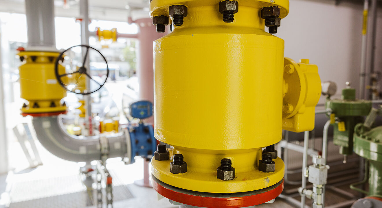 Detailaufnahme einer Rohrleitungsanlage mit großer gelber Armatur und roten Flanschmarkierungen. Die Armatur ist an graue Rohre angeschlossen, im Hintergrund sind weitere Leitungen, Ventile und technische Komponenten sichtbar.