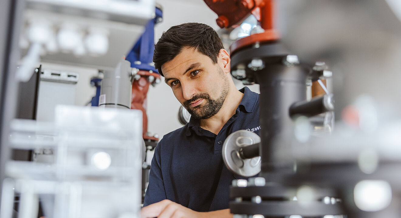 Techniker arbeitet an einer komplexen Rohrleitungs- und Ventilanlage in einem industriellen Umfeld, umgeben von schwarzen, roten und blauen Armaturen sowie metallischen Komponenten.