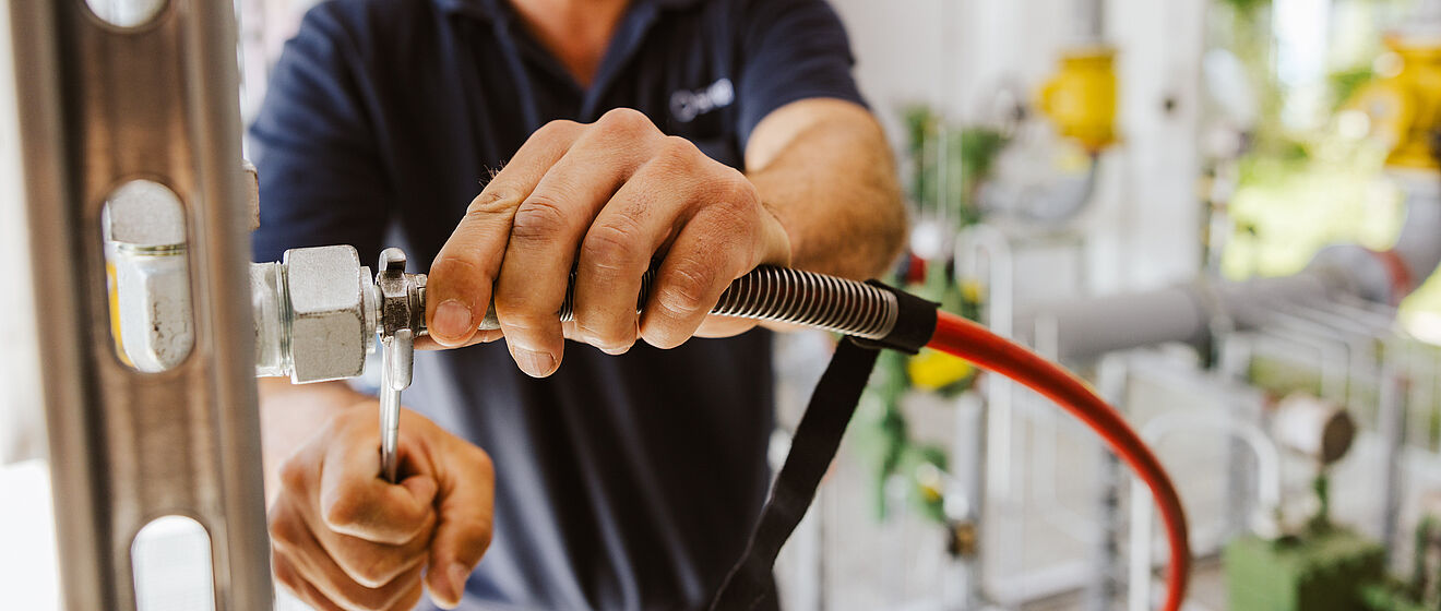Eine Person arbeitet an einer technischen Anlage mit Rohrleitungen und Armaturen. Die Person hält einen Schraubenschlüssel und zieht eine Verbindung an einem Metallrohr fest. Ein flexibler Schlauch mit rotem Kabel ist ebenfalls angeschlossen. Im Hintergrund sind weitere Rohre, Ventile und technische Komponenten sichtbar, die zu einer Versorgungsinstallation gehören. Die Szene zeigt Wartungs- oder Montagearbeiten in einem industriellen oder energietechnischen Umfeld.