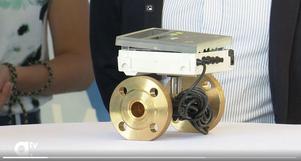 Auf einem Tisch liegt ein Messgerät, das auf einem metallischen Durchfluss-Sensor montiert ist. Der Sensor besteht aus zwei massiven, goldfarbenen Flanschen, die durch ein Rohr verbunden sind. Auf dem Sensor ist ein rechteckiges elektronisches Gerät mit Display und Bedienelementen befestigt, daneben sind Kabel sichtbar. Im Hintergrund sind Personen teilweise erkennbar, aber unscharf. Das Gerät dient zur präzisen Messung von Durchflussmengen in Rohrleitungen, typischerweise für Wasser- oder Energieanwendungen.