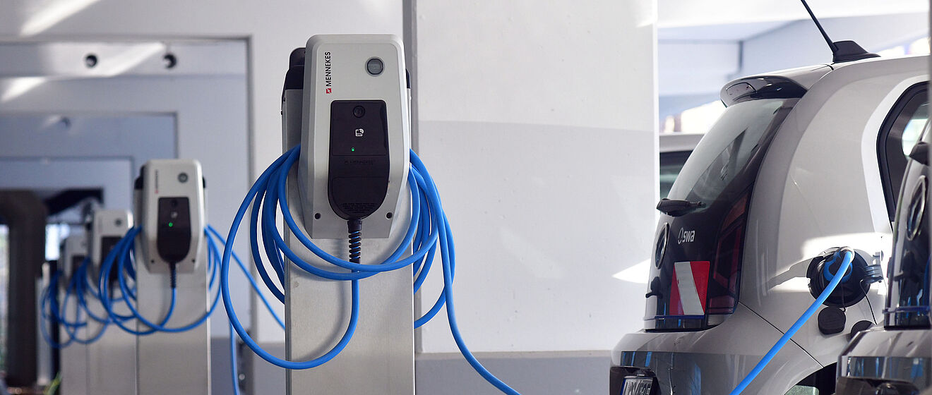 Mehrere Ladestationen für Elektrofahrzeuge in einer überdachten Ladezone: Die weißen Säulen sind mit blauen Ladekabeln ausgestattet, die ordentlich aufgewickelt sind. Rechts im Bild sind zwei Elektroautos an die Ladestationen angeschlossen. Der Boden ist teilweise mit Laub bedeckt, die Umgebung wirkt modern und funktional. Die Szene symbolisiert Ladeinfrastruktur für Elektromobilität.