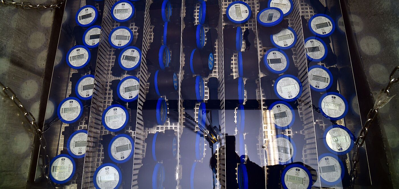Mehrere blaue digitale Wasserzähler sind in einem Metallrahmen in Reihen angeordnet. Die Zähler zeigen jeweils ein Display mit technischen Angaben und sind durch Trennwände voneinander getrennt. Die Installation befindet sich in einem rechteckigen Schacht mit Metallketten an den Seiten.