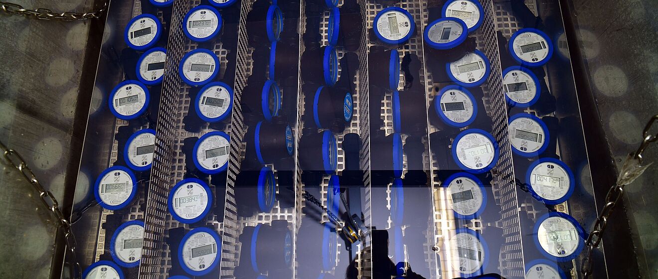 Mehrere blaue digitale Wasserzähler sind in einem Metallrahmen in Reihen angeordnet. Die Zähler zeigen jeweils ein Display mit technischen Angaben und sind durch Trennwände voneinander getrennt. Die Installation befindet sich in einem rechteckigen Schacht mit Metallketten an den Seiten.
