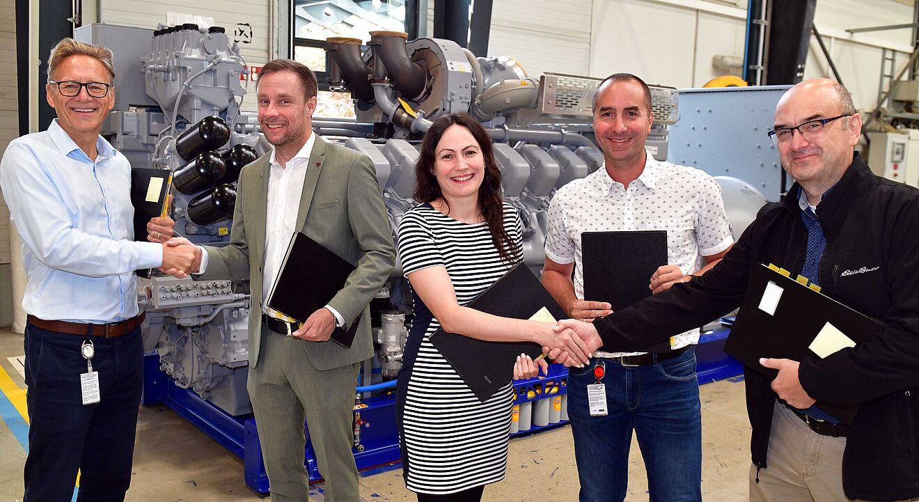 Fünf Personen stehen in einer Industriehalle vor einer großen Maschine mit grauen Komponenten und blauen Rahmen. Zwei Personen auf der linken Seite und zwei auf der rechten Seite geben sich jeweils die Hand. Alle halten schwarze Mappen in der anderen Hand. Im Hintergrund sind Teile der Maschine, Rohrleitungen und industrielle Ausstattung sichtbar. Verlinkung zur Kooperation von swa und Rolls-Royce Seite