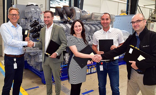 Fünf Personen stehen in einer Industriehalle vor einer großen Maschine mit grauen Komponenten und blauen Rahmen. Zwei Personen auf der linken Seite und zwei auf der rechten Seite geben sich jeweils die Hand. Alle halten schwarze Mappen in der anderen Hand. Im Hintergrund sind Teile der Maschine, Rohrleitungen und industrielle Ausstattung sichtbar. Verlinkung zur Kooperation von swa und Rolls-Royce Seite