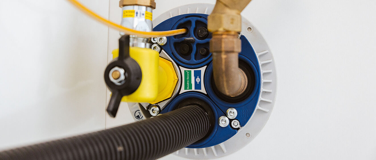 Nahaufnahme eines Hausanschlusses für Wasserleitungen: Sichtbar sind verschiedene Rohrverbindungen aus Kupfer und Messing, ein gelber Absperrhahn, ein schwarzer Schlauch sowie eine blaue Dichtungsplatte mit Markierungen. Die Installation befindet sich an einer weißen Wand. Verlinkung zur Hausanschluss Seite.
