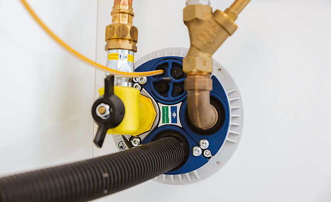 Nahaufnahme eines Hausanschlusses für Wasserleitungen: Sichtbar sind verschiedene Rohrverbindungen aus Kupfer und Messing, ein gelber Absperrhahn, ein schwarzer Schlauch sowie eine blaue Dichtungsplatte mit Markierungen. Die Installation befindet sich an einer weißen Wand. Weiterverlinkung zur swa Netze Hausanschluss -Seite 