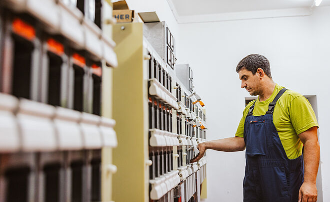 Person in Arbeitskleidung bedient Schaltanlage: Die Person trägt ein grünes T-Shirt und eine blaue Latzhose und steht vor einer Reihe von elektrischen Schaltkästen in einem Technikraum. Die Hand greift an einen der Schalter oder Hebel. Im Hintergrund sind weitere Schaltfelder und eine helle Wand sichtbar. Die Szene zeigt Arbeiten an elektrischen Anlagen in einem Innenbereich. Verlinkung zur Kabelabschaltung Seite.