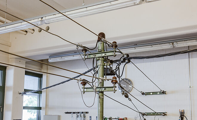 Technischer Schulungsraum mit Oberleitungsmodell: In der Mitte steht ein grüner Metallmast mit mehreren Isolatoren und daran befestigten Stromleitungen, die die Struktur einer Bahnstrom-Oberleitung simulieren. Im Hintergrund sind Werkzeuge, Kabelrollen und Sicherheitswarnschilder an der Wand angebracht. Die Szene zeigt eine Ausbildungsumgebung für Arbeiten an elektrischen Leitungen und Infrastruktur. Verlinkung zur Freileitungsisolierung Seite.