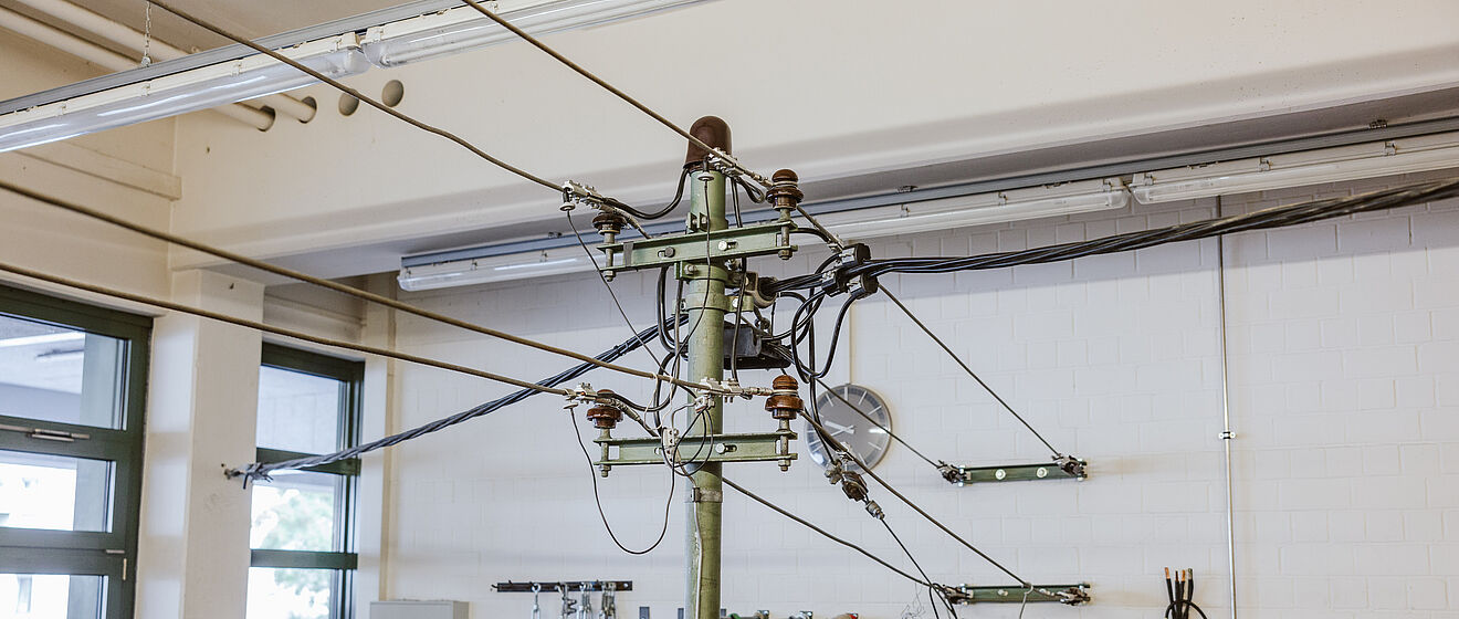 Technischer Schulungsraum mit Oberleitungsmodell: In der Mitte steht ein grüner Metallmast mit mehreren Isolatoren und daran befestigten Stromleitungen, die die Struktur einer Bahnstrom-Oberleitung simulieren. Im Hintergrund sind Werkzeuge, Kabelrollen und Sicherheitswarnschilder an der Wand angebracht. Die Szene zeigt eine Ausbildungsumgebung für Arbeiten an elektrischen Leitungen und Infrastruktur. Verlinkung zur Freileitungsisolierung Seite.