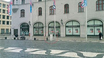 Historisches Geschäftsgebäude mit mehreren Filialfenstern der Stadtsparkasse an einem gepflasterten Stadtplatz. Vor dem Gebäude stehen vier Fahnenmasten, Sitzgelegenheiten und Poller. In der Bildmitte ist ein heller zylindrischer Pfosten am Boden rot markiert.