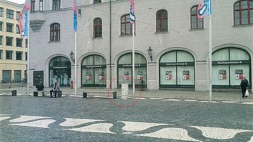 Historisches Geschäftsgebäude mit mehreren Filialfenstern der Stadtsparkasse an einem gepflasterten Stadtplatz. Vor dem Gebäude stehen vier Fahnenmasten, Sitzgelegenheiten und Poller. In der Bildmitte ist ein heller zylindrischer Pfosten am Boden rot markiert.