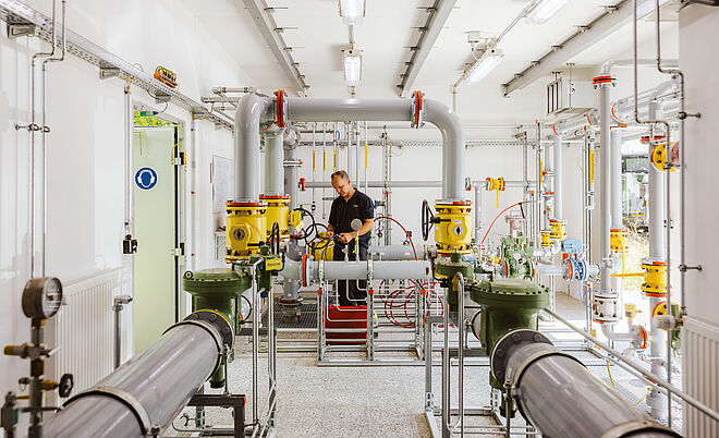 Technikraum mit komplexer Rohrleitungsanlage: Mehrere graue und gelbe Rohre, Ventile und Messgeräte sind sichtbar. Eine Person arbeitet an der zentralen Armatur, umgeben von roten Schläuchen und metallischen Verbindungen. Im Hintergrund sind Kabel, Schaltkästen und helle Beleuchtung erkennbar. Verlinkung zur Gas Seite.