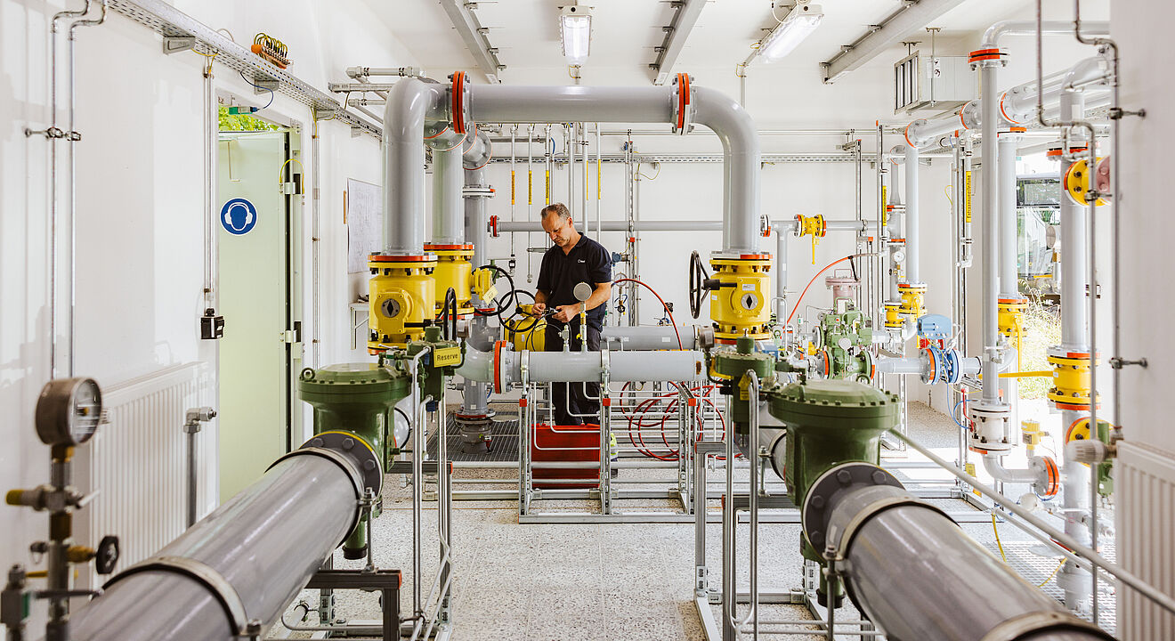Technikraum mit komplexer Rohrleitungsanlage: Mehrere graue und gelbe Rohre, Ventile und Messgeräte sind sichtbar. Eine Person arbeitet an der zentralen Armatur, umgeben von roten Schläuchen und metallischen Verbindungen. Im Hintergrund sind Kabel, Schaltkästen und helle Beleuchtung erkennbar. Verlinkung zur Gas Seite.