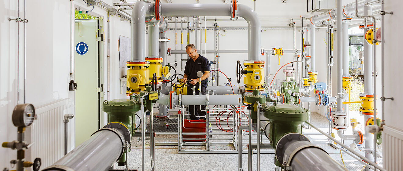 Technikraum mit komplexer Rohrleitungsanlage: Mehrere graue und gelbe Rohre, Ventile und Messgeräte sind sichtbar. Eine Person arbeitet an der zentralen Armatur, umgeben von roten Schläuchen und metallischen Verbindungen. Im Hintergrund sind Kabel, Schaltkästen und helle Beleuchtung erkennbar. Verlinkung zur Gas Seite.