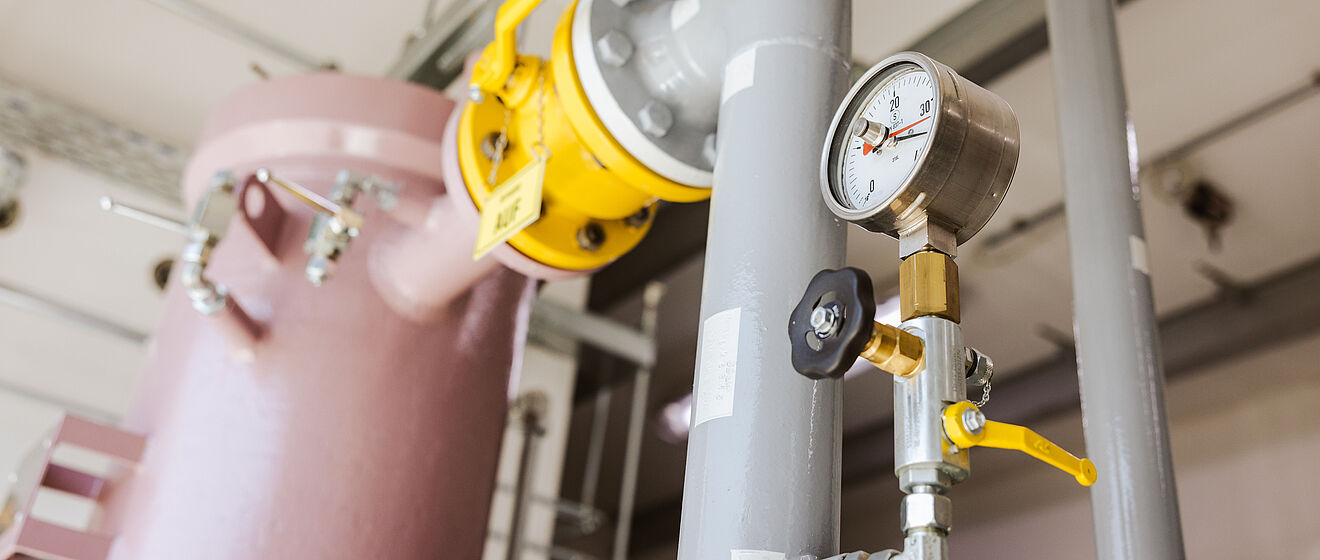 Technikraum mit Rohrleitungsanlage: Im Vordergrund ein graues Rohr mit Manometer (0–30 bar) und Ventil, daneben eine gelbe Armatur mit Hebel in geöffneter Position. Im Hintergrund ein großer rosafarbener Druckbehälter sowie weitere metallische Leitungen und Befestigungen. Verlinkung zur Lastprofile Gas Seite.