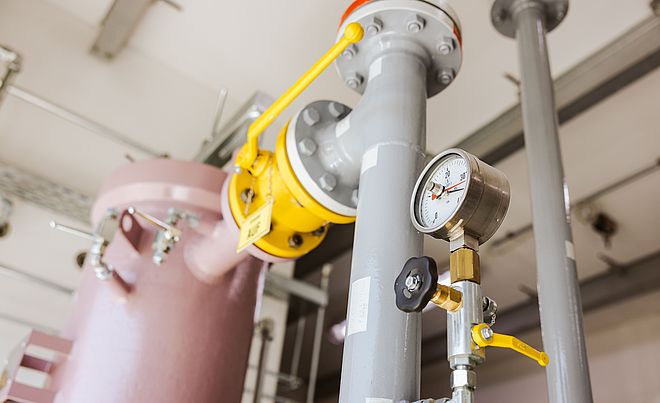 Technikraum mit Rohrleitungsanlage: Im Vordergrund ein graues Rohr mit Manometer (0–30 bar) und Ventil, daneben eine gelbe Armatur mit Hebel in geöffneter Position. Im Hintergrund ein großer rosafarbener Druckbehälter sowie weitere metallische Leitungen und Befestigungen. Verlinkung zur Lastprofile Gas Seite.