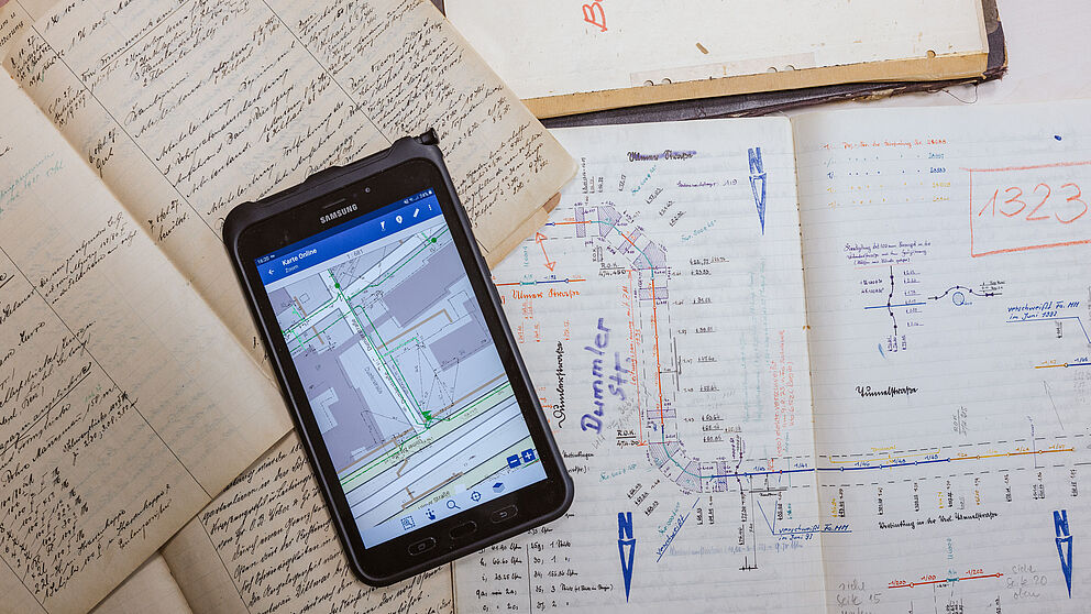 Auf einem Tisch liegen mehrere aufgeschlagene Hefte und Blätter mit handschriftlichen Notizen und technischen Zeichnungen. In der Mitte befindet sich ein Smartphone, auf dessen Bildschirm ein digitaler Lageplan mit Straßen und Leitungen angezeigt wird. Die Zeichnungen zeigen detaillierte Skizzen von Leitungsverläufen und Markierungen. Im oberen Bereich ist ein Buch mit der Aufschrift „Bau-“ zu sehen, daneben weitere technische Unterlagen.
